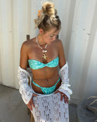 Woman in turquoise bikini and white cover-up standing against a corrugated metal wall.