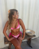 Woman in a pink bikini lying on a wooden bench against a white wall.